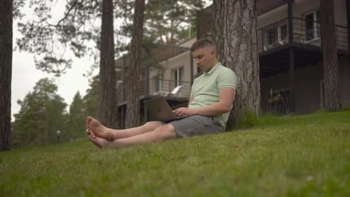 A Young Man Sits Under a Tree and Works on a Laptop A Man with a Laptop in Nature Sitting on the
