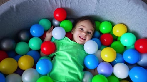 Happy smiling toddler lies in the dry pool. Charming Caucasian baby boy in a pile of colorful balls.
