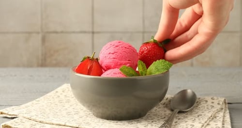 Woman adding mint to sorbet with strawberries at grey wooden table, closeup