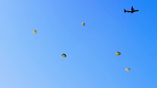 Ein Militärflugzeug setzt Fallschirmjäger frei, die unter runden Fallschirmen gegen ein hellblaues s absteigen.