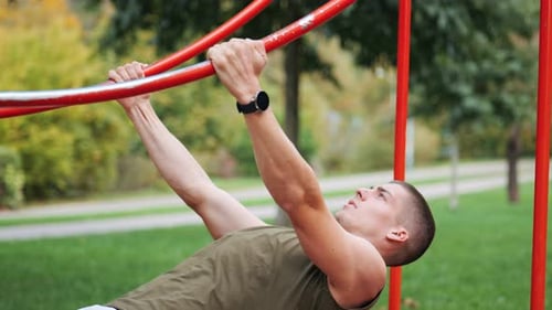 Focused young muscular athletic man doing pull-ups at red sports facility in park