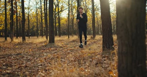 Confident Sporty Man with Curly Hair in a Black Sports Uniform Runs Through the Forest with Fallen