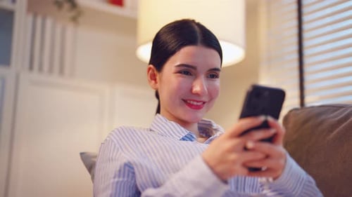 Asiatische schöne Frau sitzt auf dem Sofa, telefoniert im Haus am Handy.