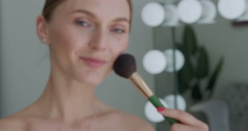 Woman Applies Makeup with Brush in Front of Mirror