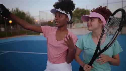 Two young women, tennis players, wearing visors and holding rackets, take selfies on a blue court af