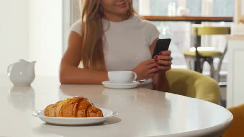 Woman Enjoying a Croissant While Using Her Smartphone at a Cozy Bakery Café