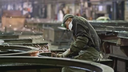 Pan shot of male worker in safety goggles using angle grinder to polish steel parts at metal