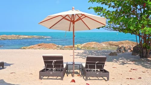 Two empty sun loungers under a large canvas shade umbrella on a white sandy beach face the rocky sho