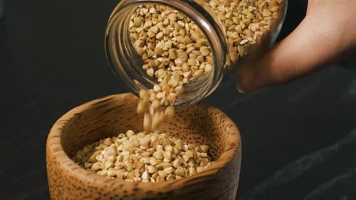 Pouring Raw Whole Grain Green Buckwheat Seeds From Glass Jar Into a Wooden Cup