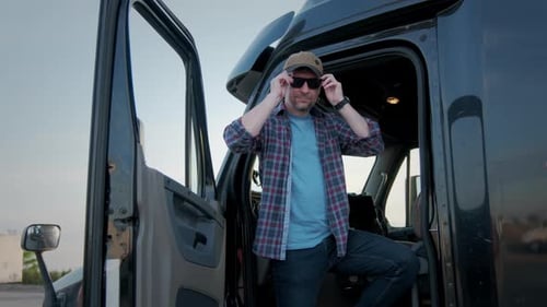 Close Up of Professional Truck Driver Sits in His Truck and Behind Him Parked Long Haul SemiTruck