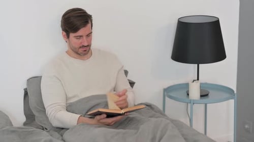 Young Man Reading Book while Sitting in Bed