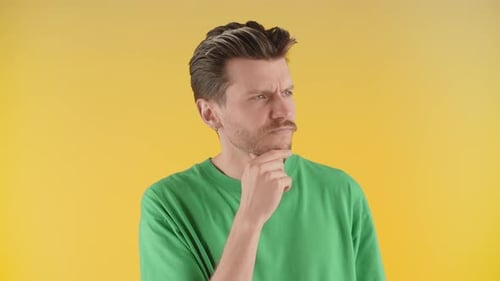 View of a Thoughtful Young Man Standing Against a Yellow Wall A Man in a Green Tshirt Rubs His Chin