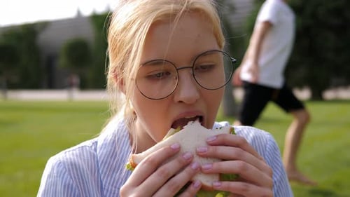 A Student Girl is Eating a Fresh Sandwich in the Park Sitting on a Green Lawn