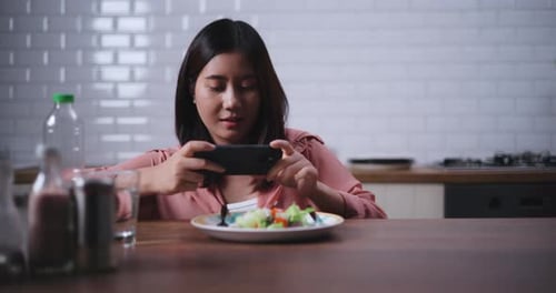 Young Asian woman sitting at a table enjoying a salad and use smartphone taking a photo in kitchen a