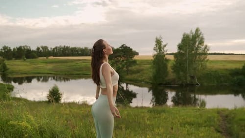 Young Woman Stretches Her Arms Up Doing Yoga Against the Background of a Pond