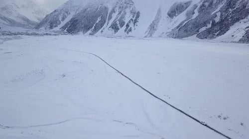 Aerial Shot of Car driving down a single snow-covered road with snowy mountains in the background