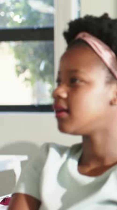 Vertical video: Panning student with pink headband listening at desk, peer raising hand, copy space