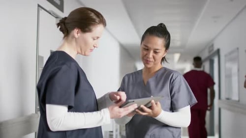 Diverse female doctors discussing work, using tablet in corridor at hospital, slow motion