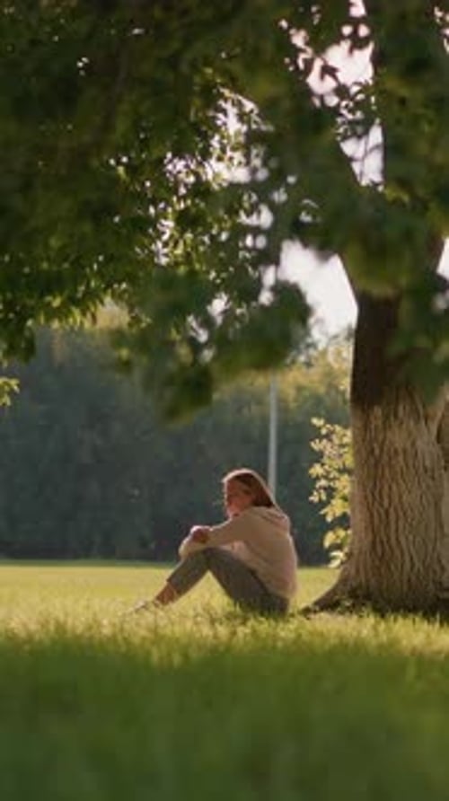 Thoughtful Woman Seated Under Tree in Sunlit Park with Greenery