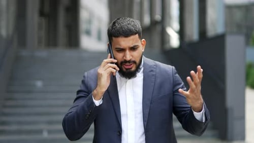 Angry adult bearded businessman arguing while talking on smartphone standing on the street near
