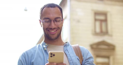 Close Up of Happy Young Handsome Smiling Male Torist with Backpack Texting Sms on Mobile Phone and