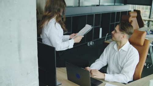 In a Modern Office a Woman Sits on a Desk with Papers Smiling and Chatting Playfully with a Male
