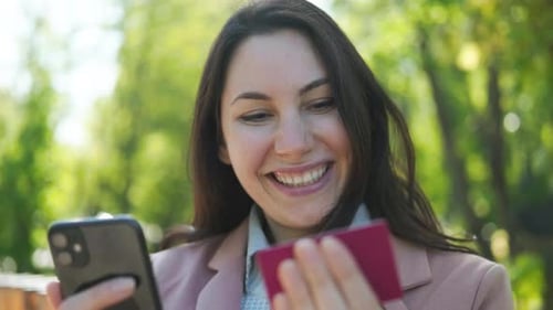 A Woman Holds a Phone and a Bank Card in Hands and is Happy About the Purchase