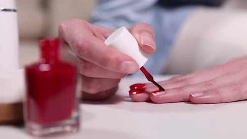 Woman applying red nail polish onto her nails at white table indoors, closeup