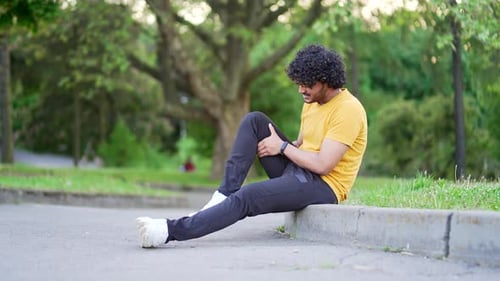 Young athlete runner sitting with muscle pain in city park. Man massaging stretching, trauma injury