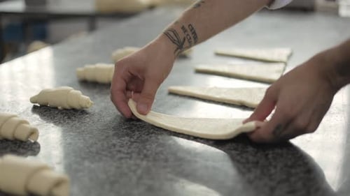 A baker forms a croissant from puff pastry at a bread factory. Close-up of male hands with tattoos.