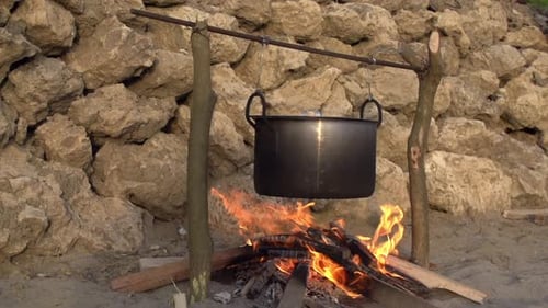 Wide shot of a metal cooking pot hanging over an open fire on a sandy beach. Flames licking up on th