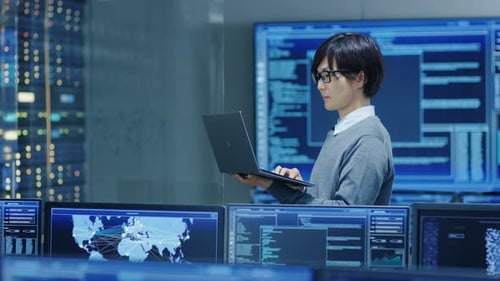 Na sala de controle do sistema, o técnico de TI trabalha em um laptop, em segundo plano, vários monitores com Gr