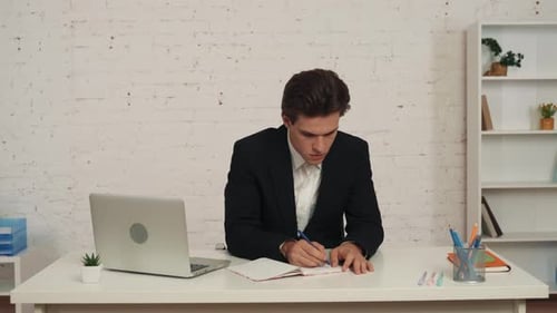 Vidéo moyenne d'un jeune homme assis à son bureau, il pense à une idée et écrit quelque chose