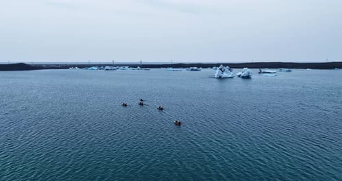 Aerial orbit of kayakers paddling across a cold blue glacial lagoon near Vatnajökull, Iceland, with