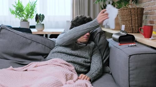 Sick Young Guy Coughing Sneezing Into a Nasal Tissue Sitting on Couch at Home