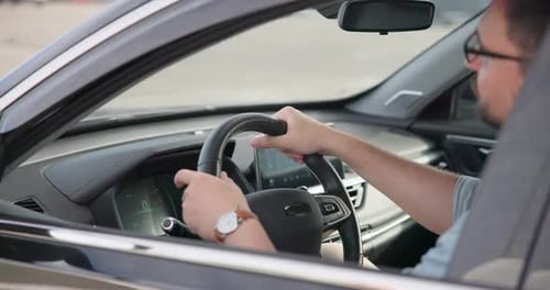 Young Caucasian Man Sits in a Car on a Parking Lot Puts Hands on a Steering Wheel