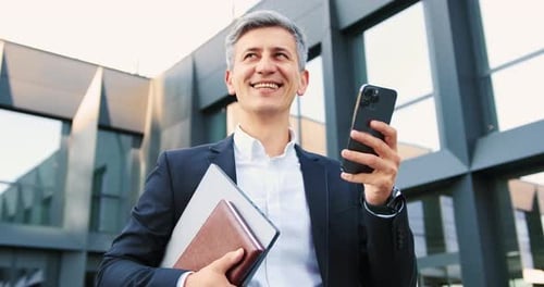 Smiling Businessman Uses Smartphone Outside Modern Office Building