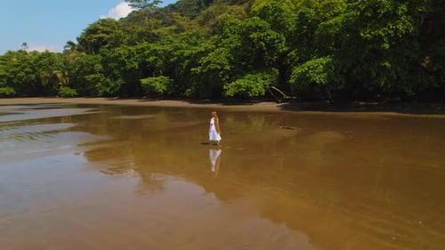 Young woman in white dress walks on wet sand beach with lush jungle on shore in Costa Rica