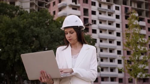 A Female Engineer in a White Helmet with a Laptop at a Construction Site