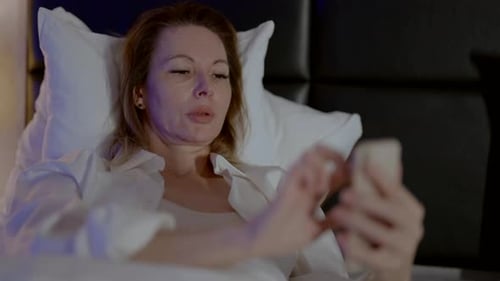 An Adult Woman Uses a Smartphone and Lies on the Bed at Night