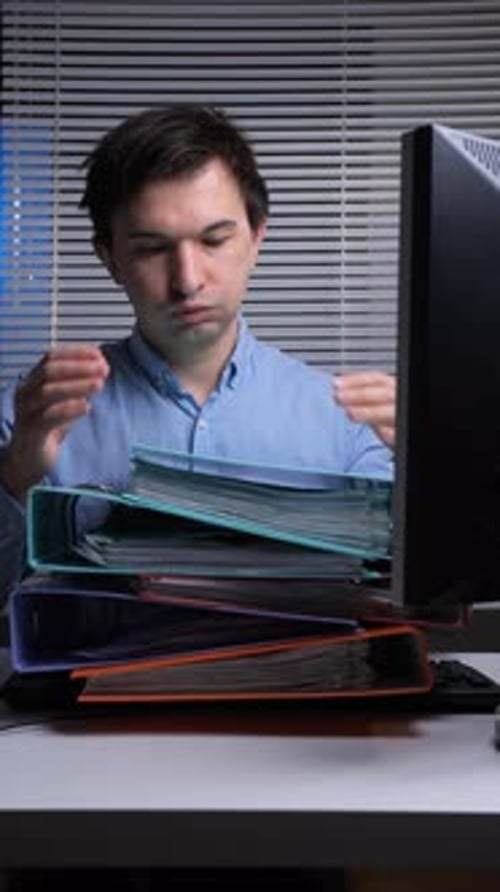 Vertical Video Young Man Standing at His Office Desk Carrying a Large Heavy Stack of Overflowing