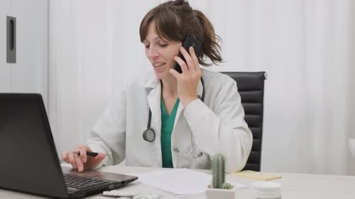 Female Physician Talking To The Phone While Browsing On Her Laptop Inside Her Clinic. wide shot