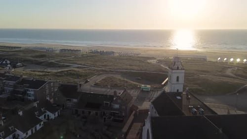 Aerial view of a seaside town at sunset, showing red-roofed houses, a historic church, sandy dunes