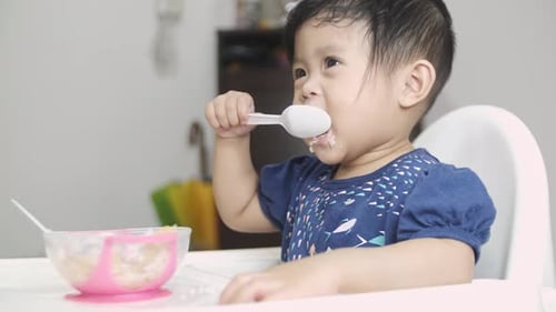Nahaufnahme eines süßen kleinen asiatischen Kindes, das auf dem Tisch sitzt und frisches Püree isst. Nahaufnahme.