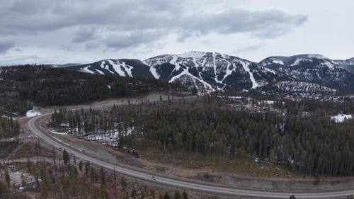Aerial view of a winding mountain road cutting through a dense coniferous forest with snow-capped