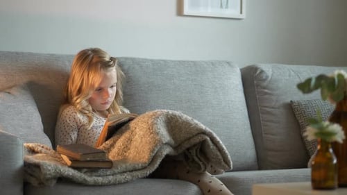 Young girl sits on couch reading a book covered by a blanket. Tilt down shot.