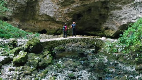 Drone view of two hikers with backpacks standing on a stone bridge.