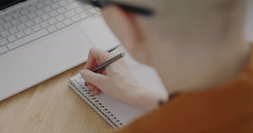 Over the Shoulder View of Female Hand Writing Information in Notebook and Using Laptop at Desk