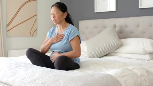 Femme asiatique âgée ressentant une gêne thoracique, plaçant les mains sur la poitrine et l'abdomen, respirant sur le lit