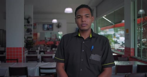 Latino waiter standing in restaurant dining area, neutral expression, hands at sides, wearing